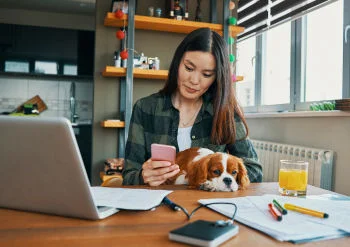 一名女士坐在桌前使用手機，正看著 LibreLink 應用程式上的葡萄糖數值，桌上擺有筆記型電腦、文件、辦公用品及柳橙汁，一隻寵物狗坐在女士腿上並將頭靠在桌上。