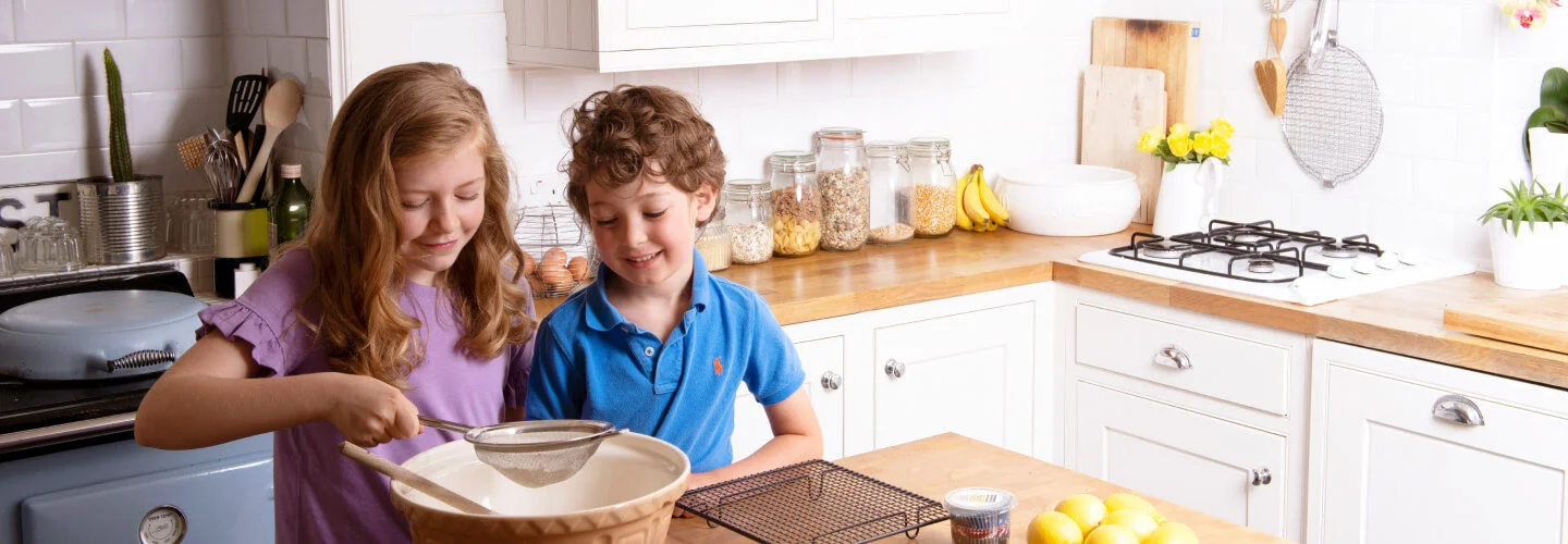 Duas crianças estão em uma cozinha bem iluminada, com armários brancos e bancadas de madeira. Uma criança está peneirando farinha em uma tigela grande enquanto a outra observa. Na bancada, há ingredientes como ovos, limões e manteiga. Ao fundo, há um fogão com panelas e prateleiras com potes de vidro.