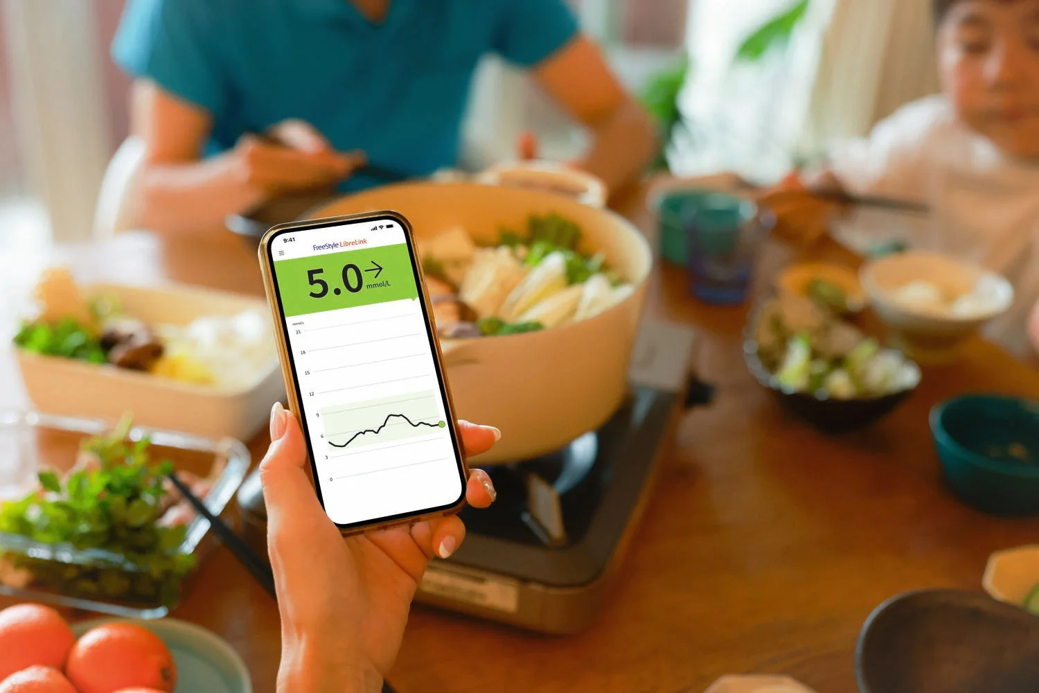 Person holding a smartphone displaying FreeStyle Libre glucose readings above a dining table with food and drinks.