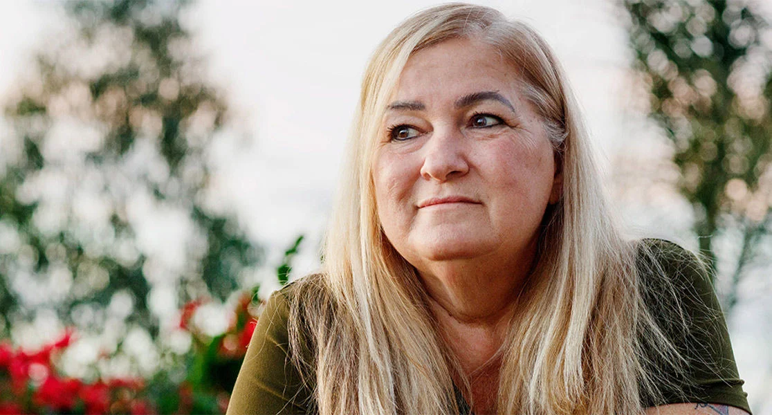 A woman with long light hair outdoors, with blurred trees and flowers in the background.