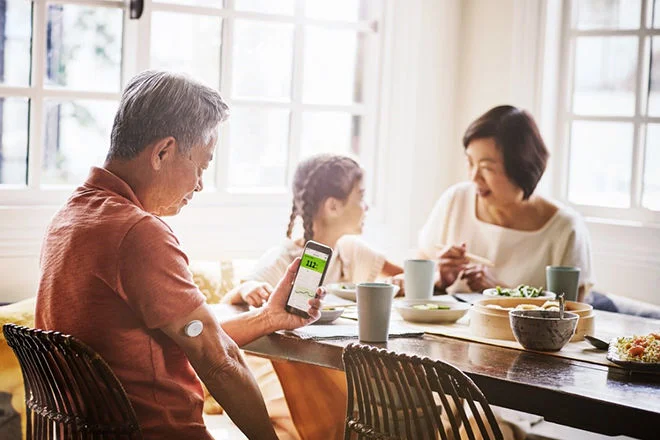 Familia sentada a mesa para sua refeição, em primeiro plano a um senhor de idade usando um sensor FreeStyle Libre e observando seus dados de glicose no aplicativo FreeStyle LibreLink