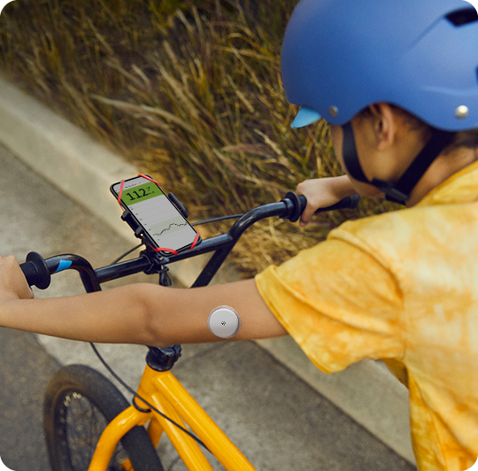 一名男孩左手臂配戴FreeStyle Libre2，頭戴安全帽正在騎腳踏車，腳踏車前方架著智慧型手機，手機螢幕上顯示LibreLink畫面。