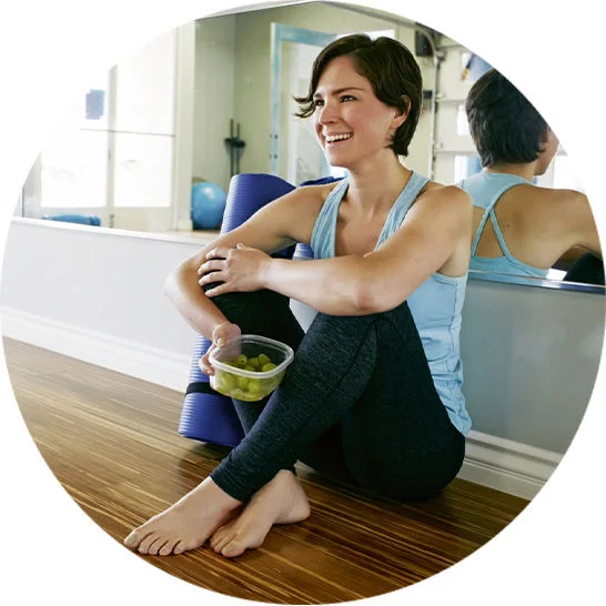 Person seated on a gym floor holding a container of grapes beside an exercise mat.