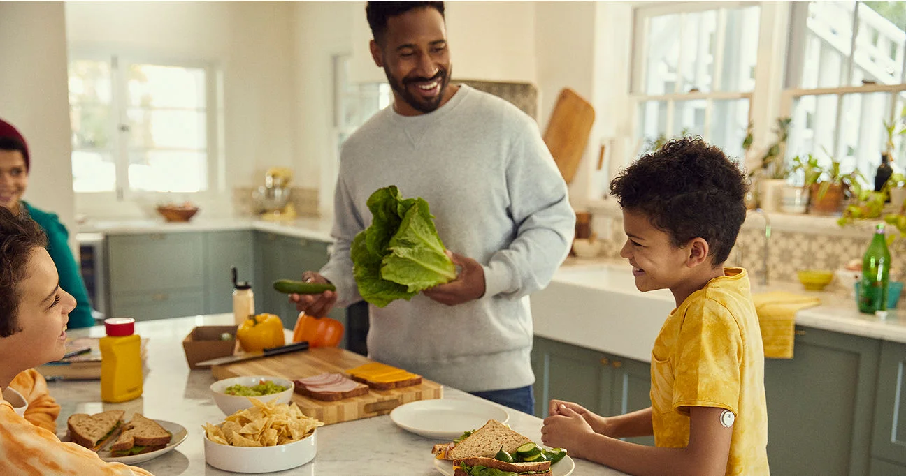 Familia reunida en la cocina durante la merienda; el padre cocina mientras su hijo, que usa el sensor FreeStyle Libre, le ayuda con alegría