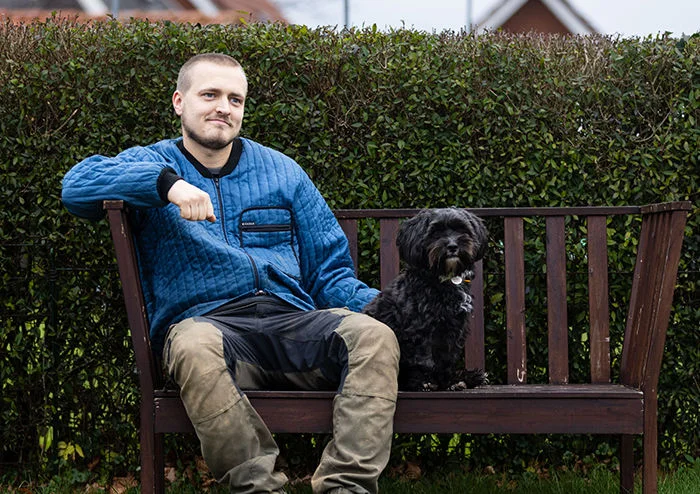 En person sidder på en bænk ved siden af en lille sort hund. Personen har ansigtet sløret.