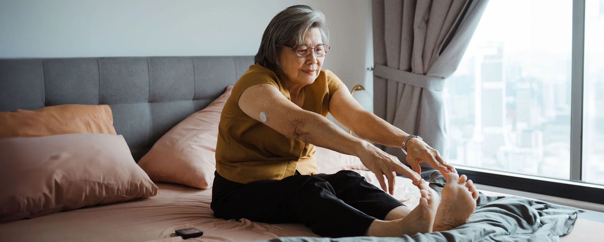 Person stretching on a bed beside a window, wearing a glucose monitor, under a “diabetes habits” article heading.