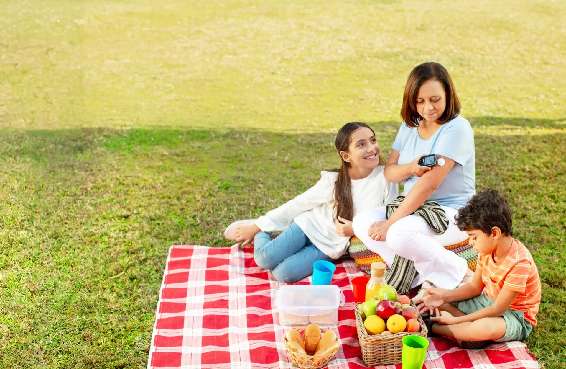 Mulher com duas crianças fazendo um piquenique, sentados sobre uma toalha xadrez com frutas e lanches, enquanto ela mede a glicose com um leitor.