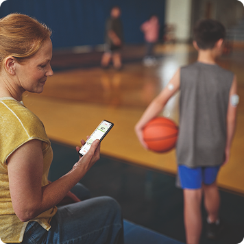 A person wearing a continuous glucose monitor (CGM) checks a smartphone while watching a child with a basketball in a gym.