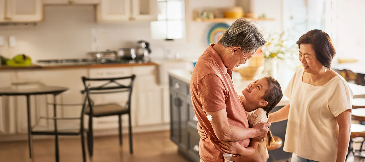 祖父手臂佩戴 FreeStyle Libre 2 傳感器與孫女擁抱歡笑，祖母站在一旁微笑注視兩人，背景為餐桌。