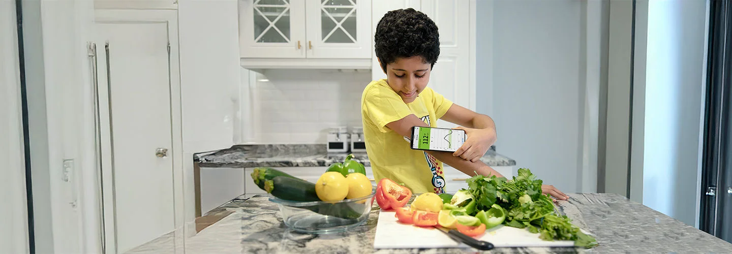 Garoto vestindo uma blusa amarela em uma cozinha com alguns legumes e vegetais enquanto escaneia seu sensor FreeStyle Libre para visualizar seus dados de glicose no aplicativo FreeStyle LibreLink