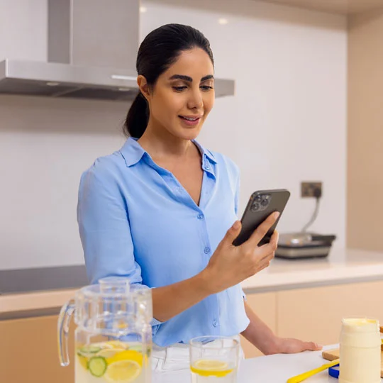 A happy woman standing in the kitchen, looking on her mobile screen.