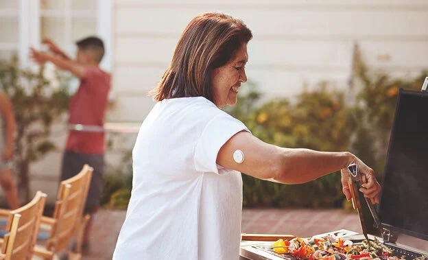 Mujer cocinando vegetales a la parrilla mientras usa el sensor FreeStyle Libre en el brazo 