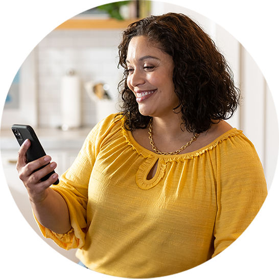 Person in a yellow blouse seated in a bright kitchen, holding and looking at a smartphone, with shelves and countertops blurred in the background.