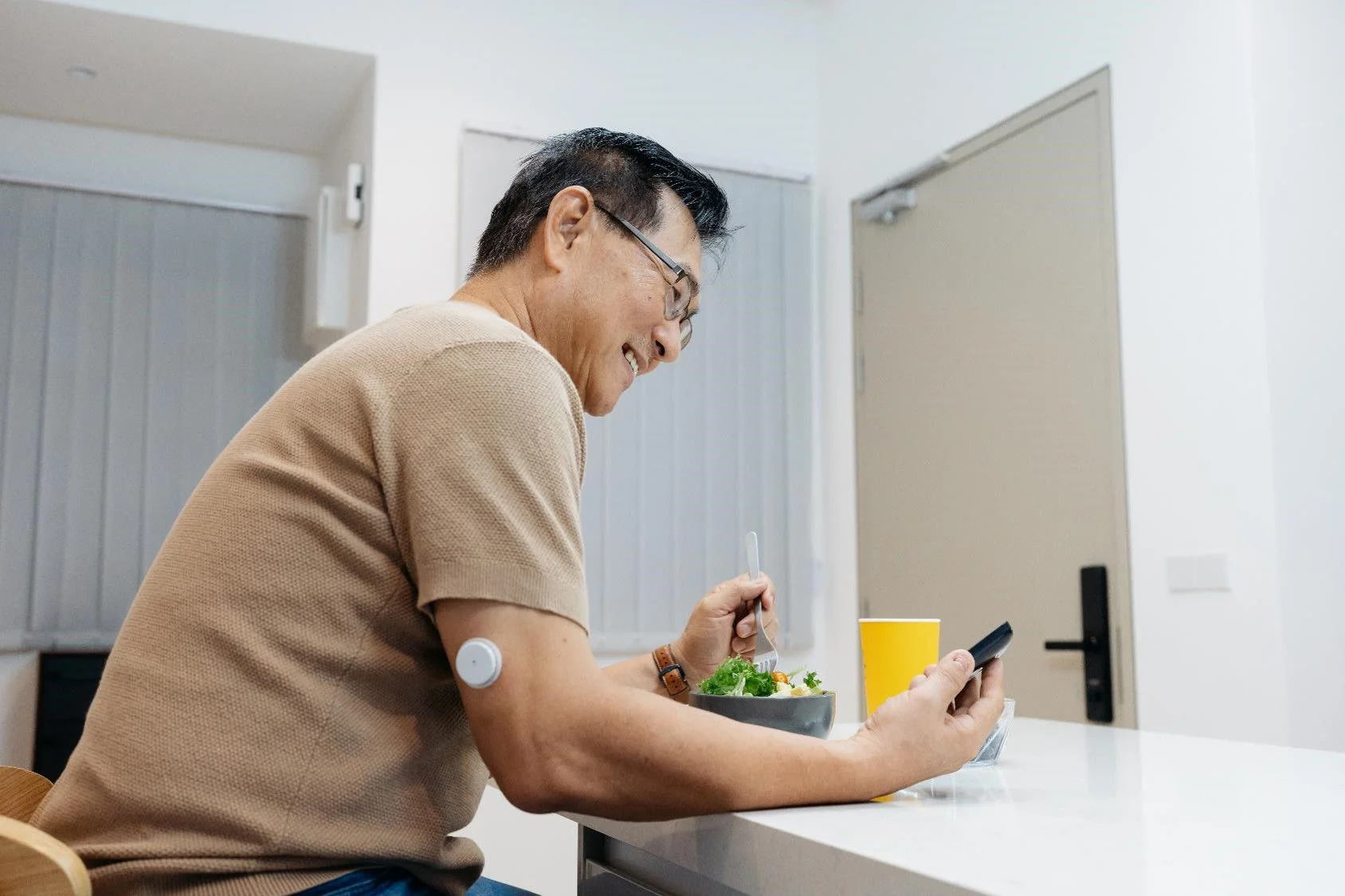 Person wearing a glucose sensor on upper arm checks phone while eating a salad at a kitchen counter.