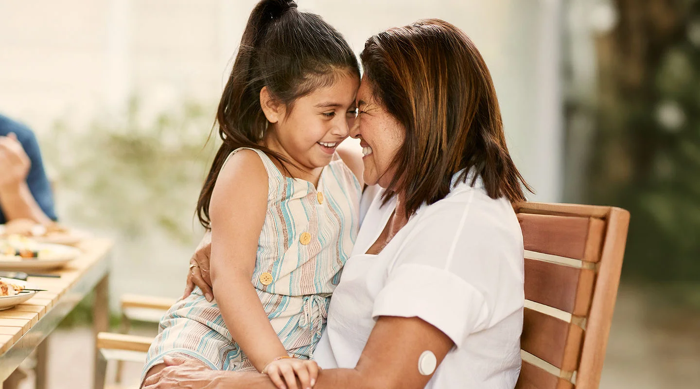 A woman sitting outdoors hugs a young girl while wearing a FreeStyle Libre sensor on her upper arm.
