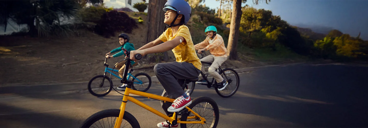 Três pessoas andando de bicicleta em uma trilha pavimentada.Há um menino andando em uma bicicleta amarela, e em seu braço há um sensor FreeStyle Libre visível, utilizado para monitoramento contínuo de glicose