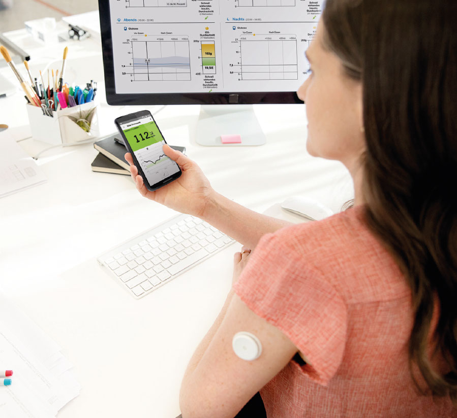 Eine Frau sitzt an einem Schreibtisch. Sie prüft ihren Glukosewert am Smartphone.