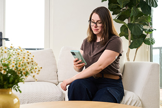 Frau sitzt auf dem Sofa und hält ein Smartphone in der Hand. Sie trägt einen Sensor am Oberarm.