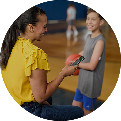 Eine Mutter überprüft die LibreLinkUp-App, während ihr Sohn, der einen FreeStyle Libre-Sensor trägt, in einer Sporthalle einen Basketball in der Hand hält.