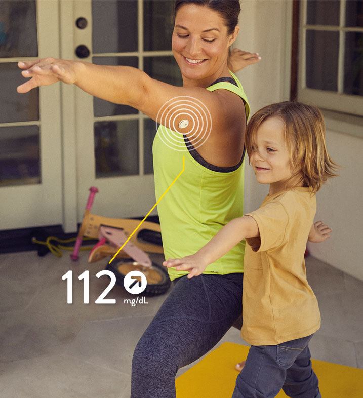 Eine Frau und ein Kind machen Yoga. Ein Smartphone mit Anzeige des Glukosewerts ist abgebildet. 