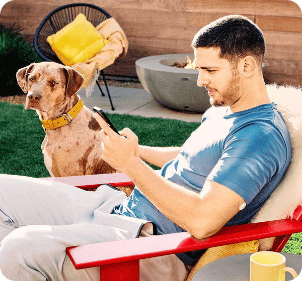 A FreeStyle Libre 3 user sitting in a garden using his phone while his dogs sits next to him, a visible FreeStyle Libre 3 sensor is applied on his arm.