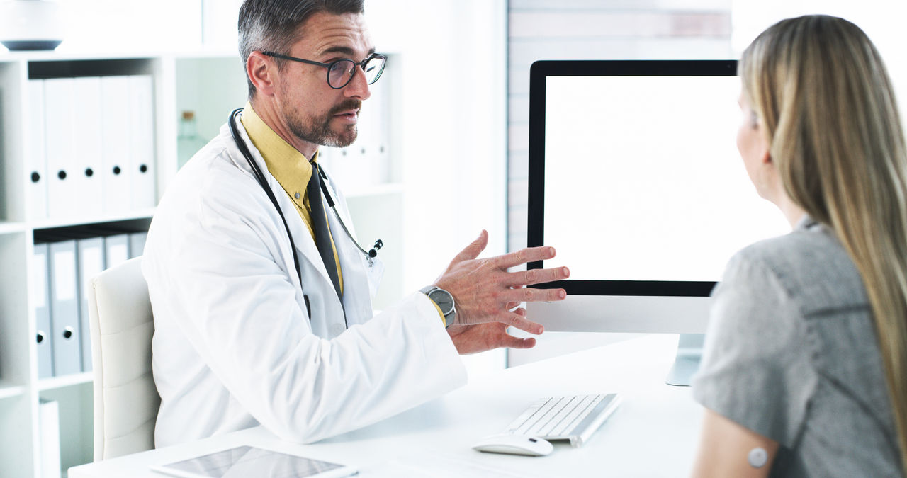 Male doctor talking to a female patient with FreeStyle Libre 3 sensor on arm . They are both sitting in the doctor's office staring at a computer