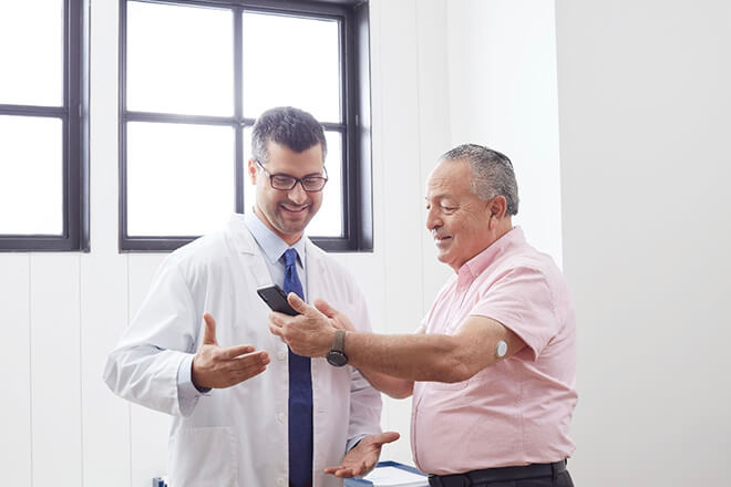 Em um consultório o paciente, vestindo uma camisa rosa e calça social preta, está utilizando o sensor FreeStyle Libre no braço esquerdo, enquanto mostra os resultados de exame no celular para o médico, a sua direita, que veste um jaleco branco, gravata azul e óculos preto.