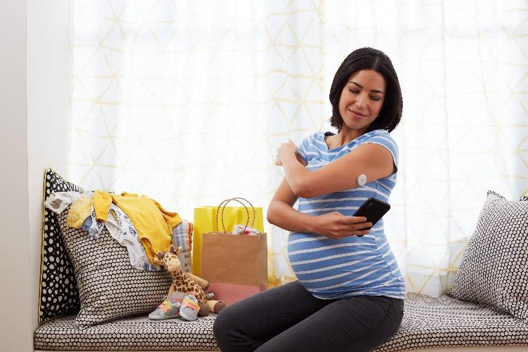 Mulher grávida apontando a tela do celular para o sensor de glicemia no seu braço esquerdo, ela está sentada em um sofá com roupas de bebê apoiadas em suas almofadas.