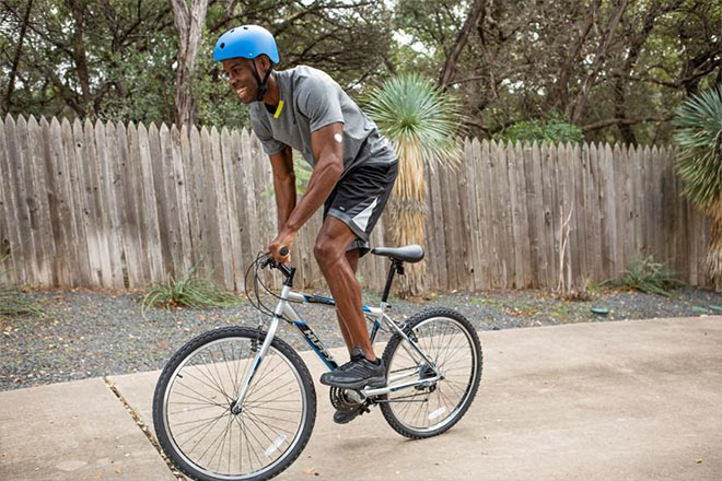 Homem andando de bicicleta usando o sensor FreeStyle Libre no braço esquerdo. O homem está andando de bicicleta em uma calçada, próximo a um muro de madeira com arvores atrás.