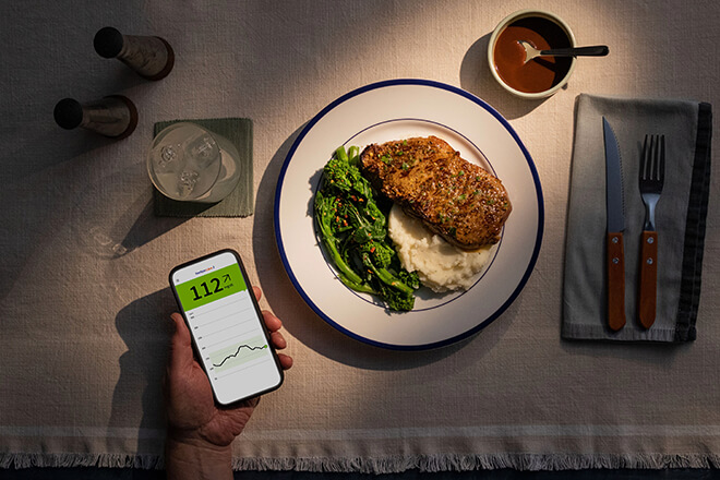 Mesa com prato com bife, legumes e puré, com utilizador monitorando níveis de glicose com aplicativo no celular.