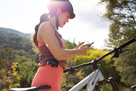 Mulher branca, de cabelos castanhos, parada ao lado de sua bicicleta, verificando o nível de glicemia pelo aplicativo da FreeStyle Libre.