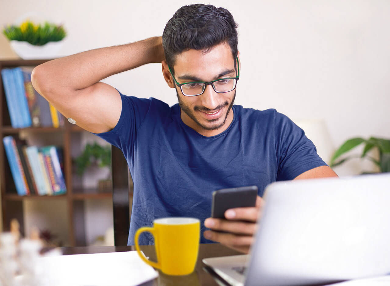 Mann sitzt am Schreibtisch mit Laptop und gelbem Becher und schaut auf sein Smartphone.