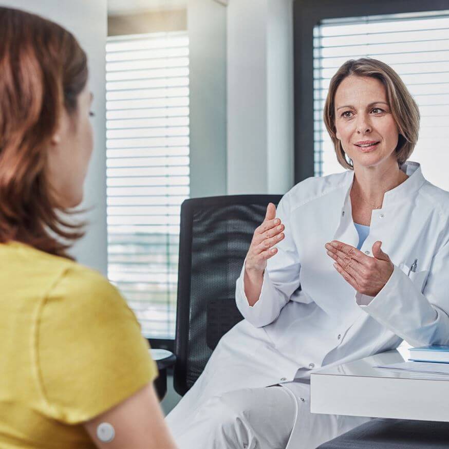 Médecin en blouse blanche discutant avec une patiente en haut jaune dans un cabinet.