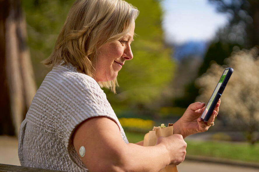 Une femme est assise sur un banc dans un parc, un smartphone à la main. Elle porte un capteur sur le haut du bras.