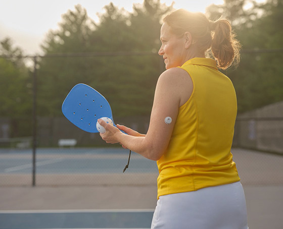 Une femme en haut sans manches jaune tenant une raquette de pickleball bleue et une balle blanche sur un court de tennis. Elle porte un capteur sur le haut du bras.