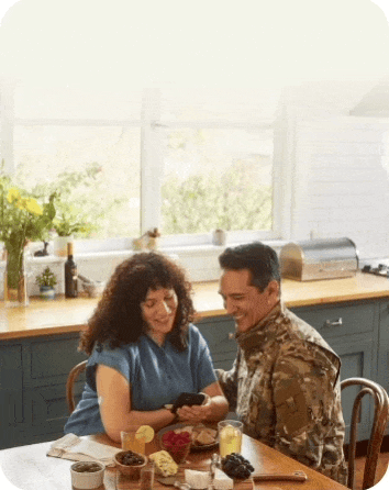 Woman checking her blood glucose on a mobile device at the breakfast table while a man in uniform looks on.