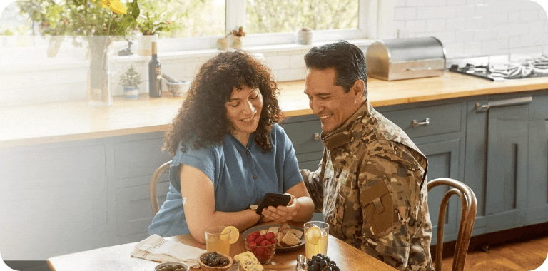 Woman checking her blood glucose on a mobile device at the breakfast table while a man in uniform looks on.
