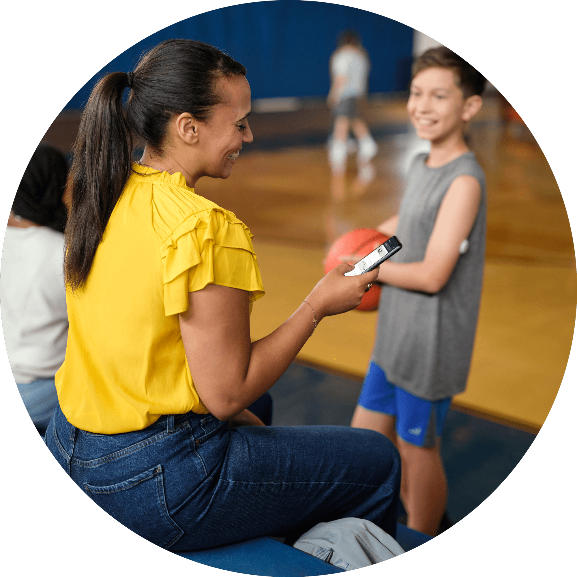 Mother checking LibreLinkUp app while her son with a FreeStyle Libre sensor holds a basketball in a gym