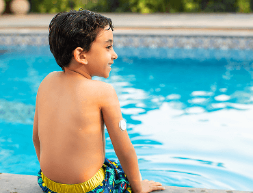 A young boy wearing a glucose sensor sitting at a pool.
