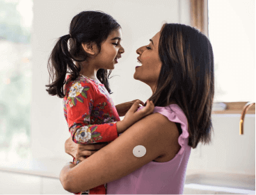 A mother wearing a glucose sensor hugging her 5 year old daughter as they smile at each other.