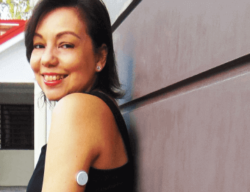 A young woman wearing her Freestyle Libre sensor leans against a wall and smiles at the camera.