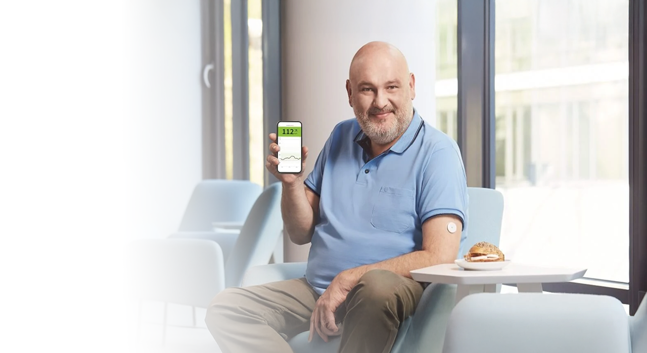 Kobieta siedzi przy kuchennym blacie, uśmiecha się sprawdzając poziom cukru w swoim telefonie, cieszy sie wygodą systemu Freestyle Libre 2 do monitorowania glikemii bez nakłuwania palców. Talerz z jedzeniem, niebieski kubek i żółte róże rozjaśniają nasłonecznione wnętrze.