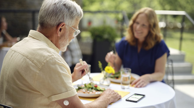 Older man wearing a continuous glucose monitor on his arm, eating a meal outdoors with a woman, with a smartphone displaying a glucose graph on the table