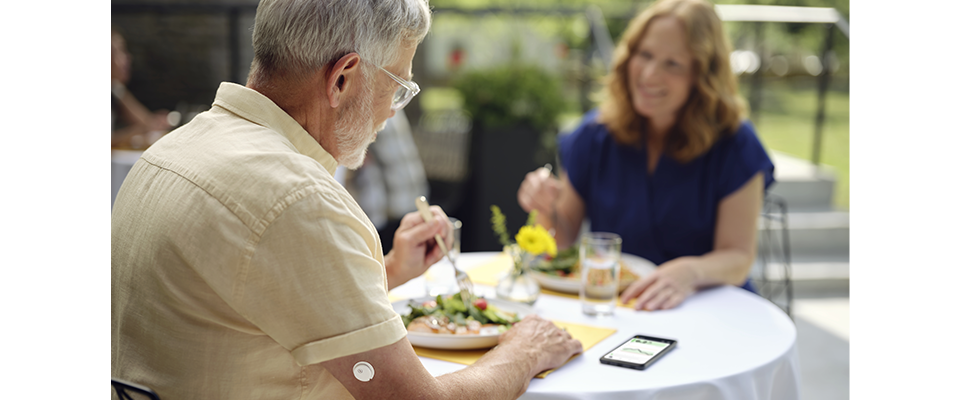 Older man wearing a continuous glucose monitor on his arm, eating a meal outdoors with a woman, with a smartphone displaying a glucose graph on the table