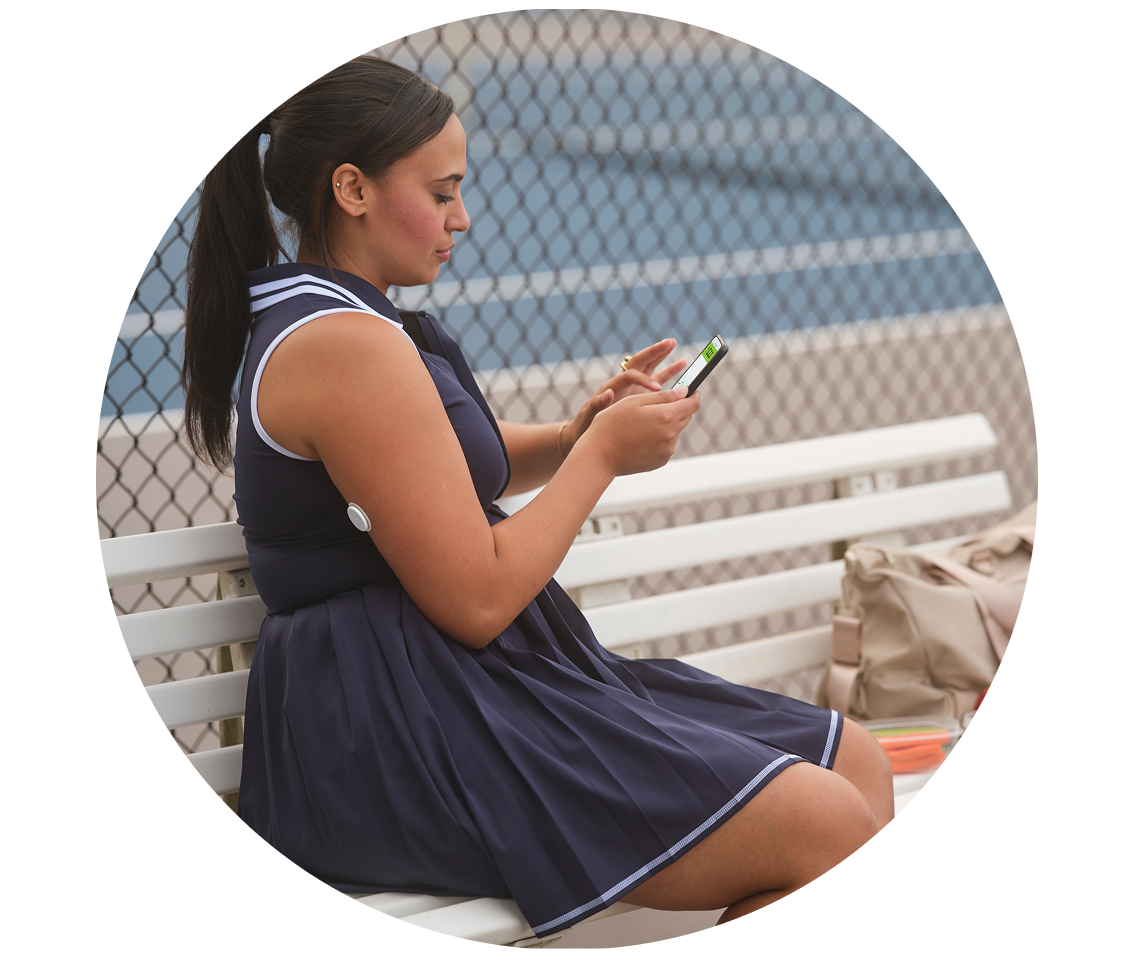 Woman in a navy tennis dress sitting on a bench beside a court, checking her phone. A FreeStyle Libre 2 Plus glucose sensor is visible on her upper arm.