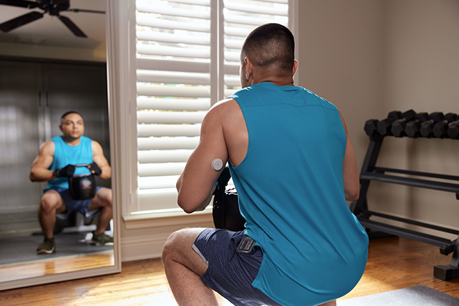 Homem usando uma camiseta azul e realizando um exercicio fisico com pesos em uma sala. Em seu braço há um sensor FreeStyle Libre
