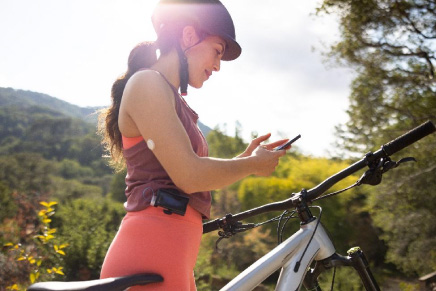 Mulher andando de bicicleta ao ar livre, enquanto verifica seu smartphone. Em seu braço há um sensor FreeStyle Libre e o dia está ensolarado