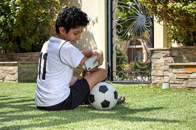 Garoto sentado na grama com uma bola de futebol, enquanto faz um sacn com seu smartphone do sensor FreeStyle Libre