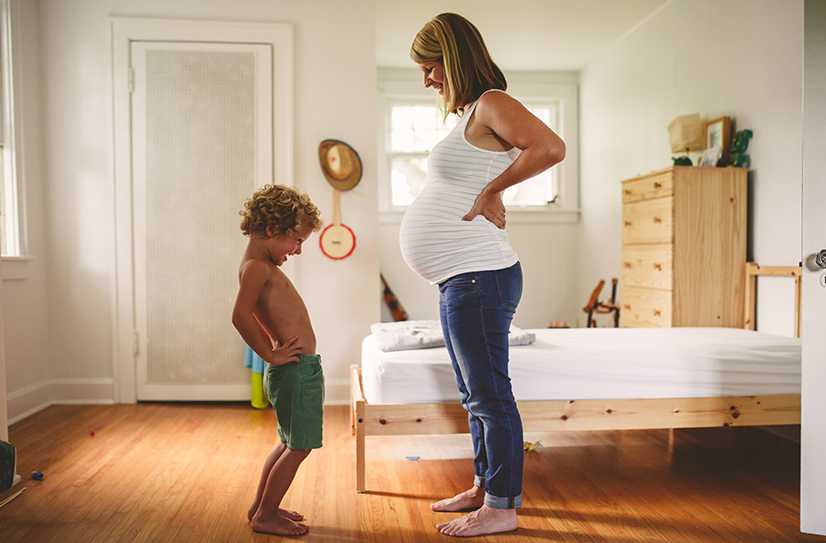 Mulher gestante com blusa branca em uma pose que evidencia sua barriga junto a seu filho, amabas estão sorrindo