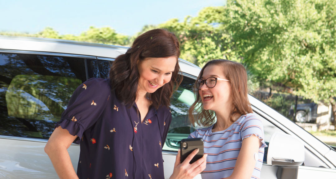  To personer smile og ser på en telefon ved siden af en bil